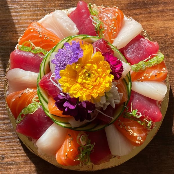 A colorful sushi cake with various fish, cucumber, and edible flowers on a wooden table.